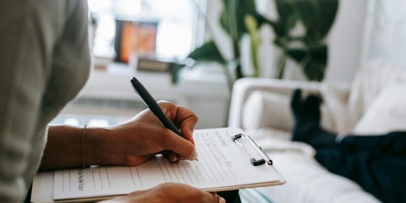 Therapist taking notes manually as her patient lies down, highlighting the lack of mental health case management software.